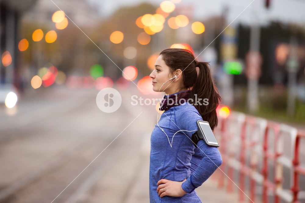 Young woman with smartphone and earphones, enjoying music while running in the city against a busy road
