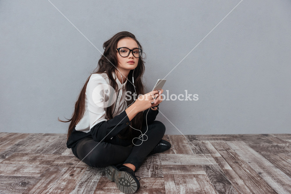 Cute woman in glasses sitting on the floor with phone and listening to music. Side view. Isolated background.