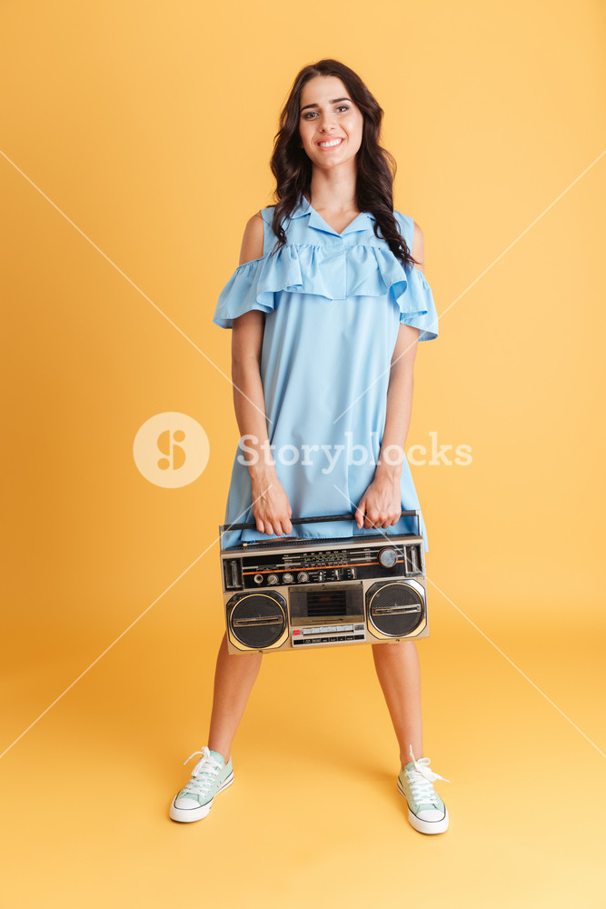 Portrait of a joyful woman in blue dress listening to music with a boombox on an orange background