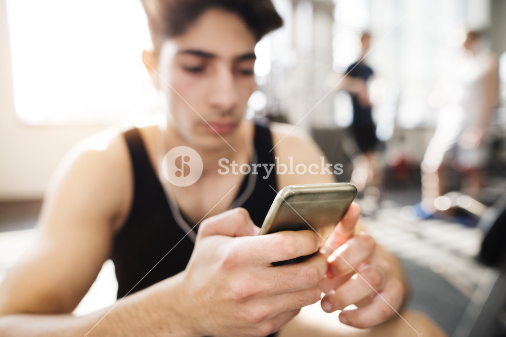 Hispanic fitness man in gym resting, holding smart phone, earphones in his ears listening music