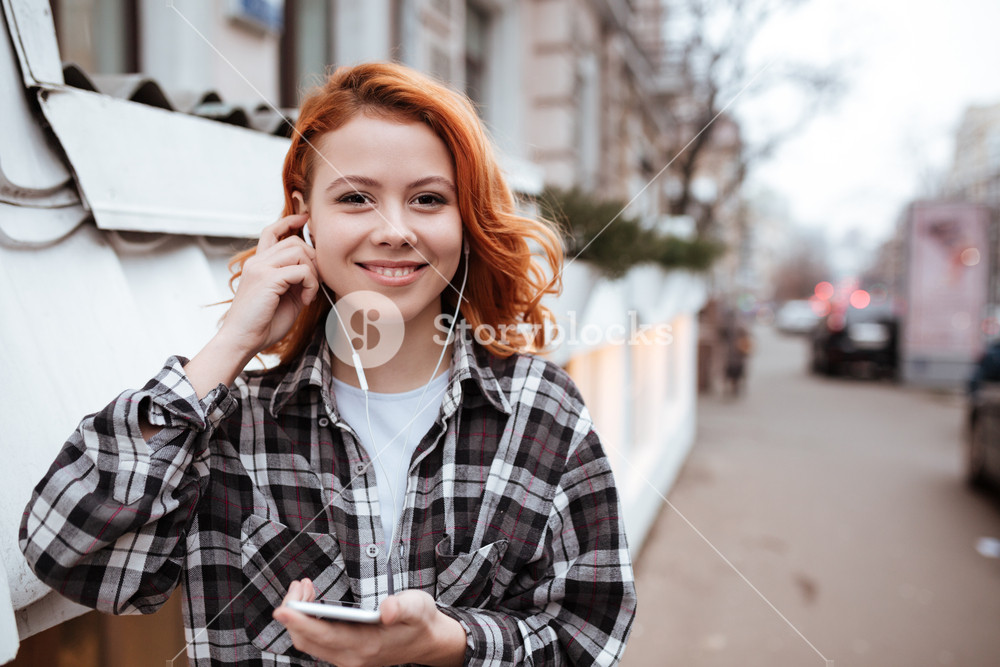 Happy young woman with red hair walking outdoors, listening to music and holding phone.