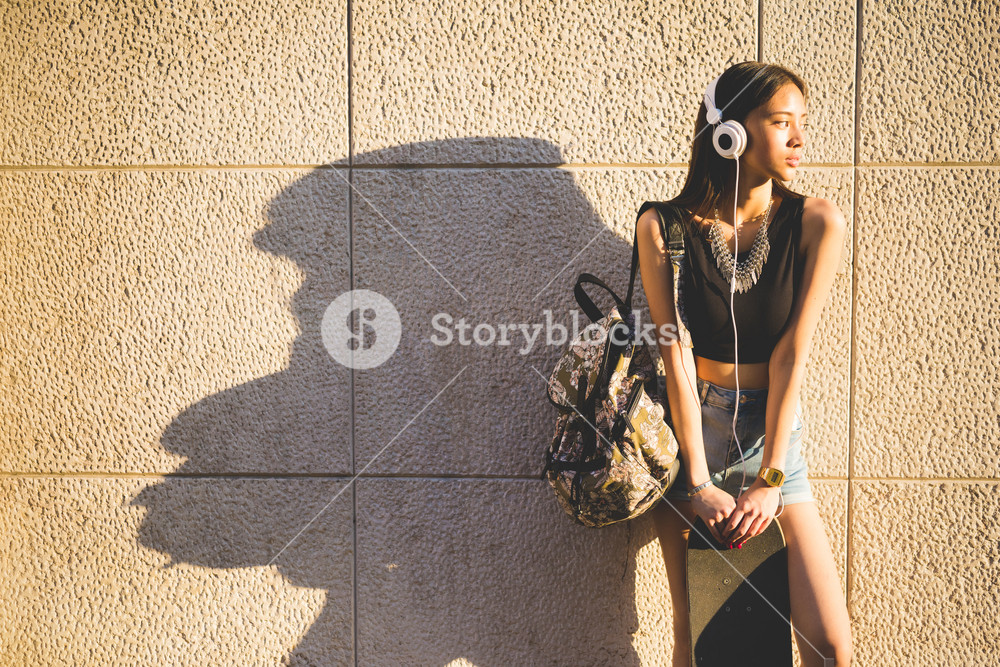 Young woman listening to music with headphones, holding a backpack and a skateboard - technology, sportive concept