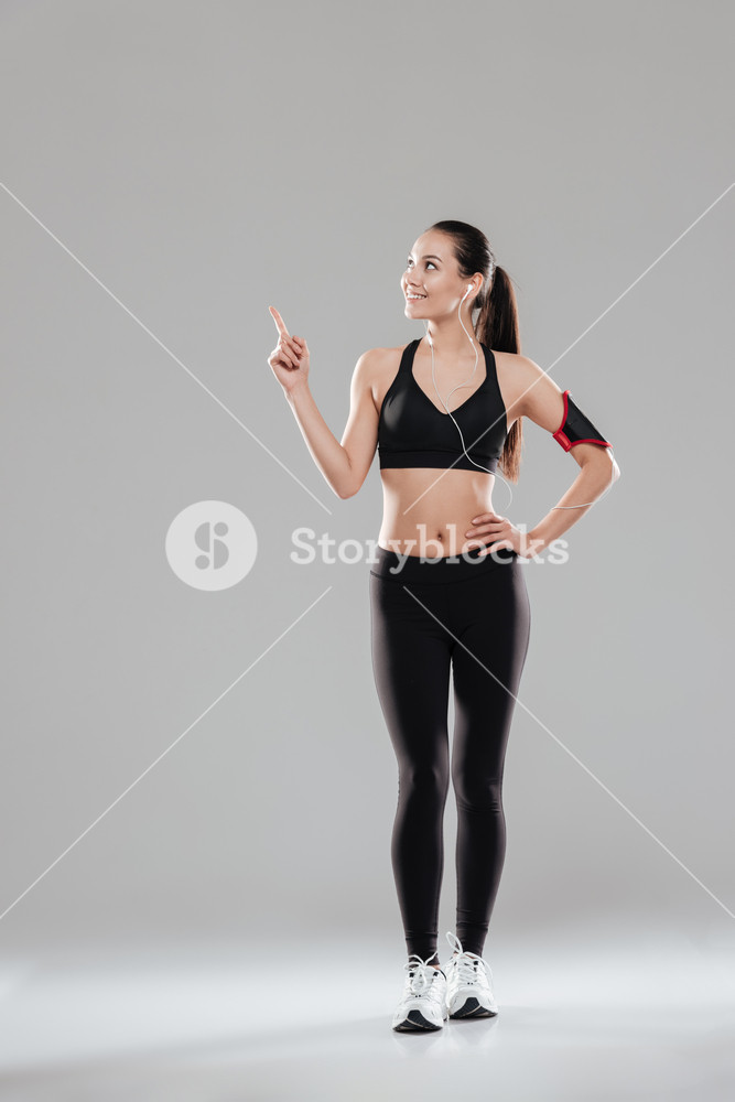 Photo of joyful young woman standing over grey background listening to music with earphones while pointing.