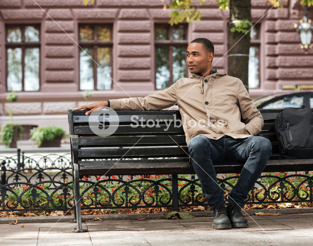 Picture of african happy man sitting on wooden bench near backpack ...