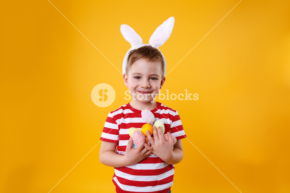 Portrait of a joyful young child wearing bunny ears and holding a bunch of colorful Easter eggs isolated on an orange background