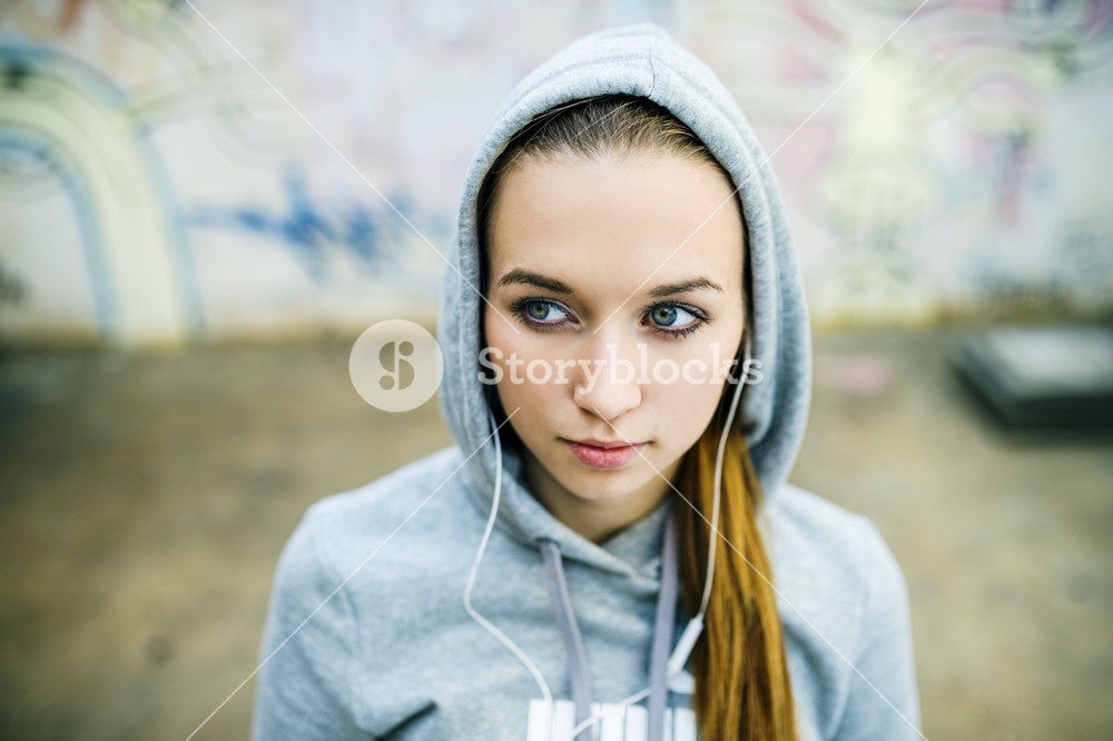 Serious teenage girl with hood on listening to music from headphones standing on background of graffiti wall