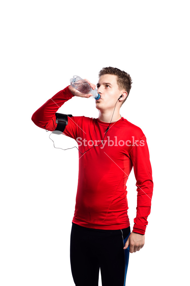 Young male athlete in blue sports sweatshirt, using smartphone on his arm, hydrating with water bottle. Attractive sportsman with earphones, enjoying music. Studio shot on white background, isolated.