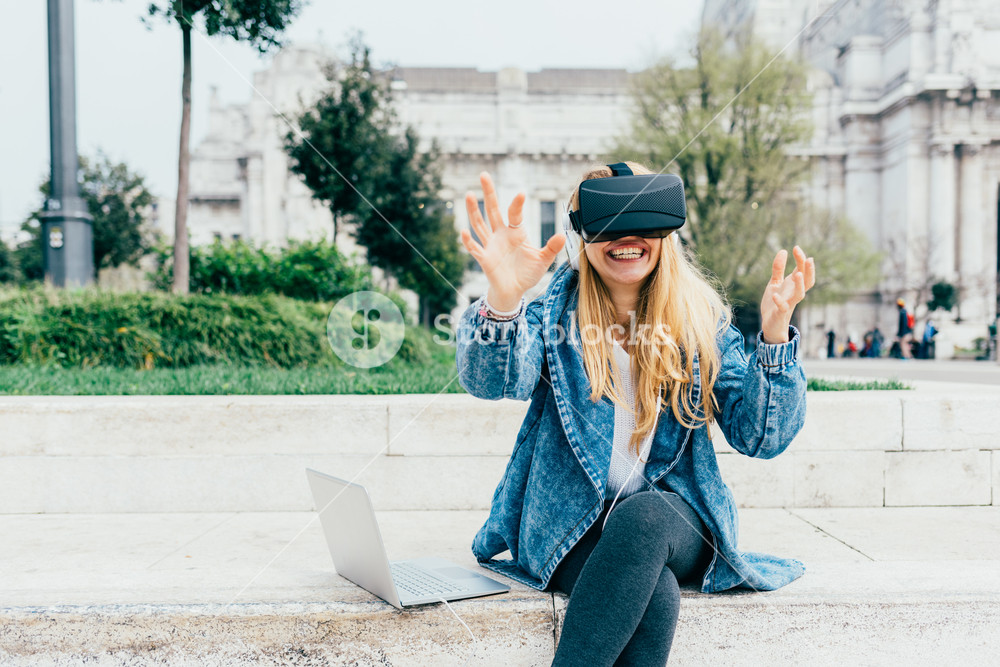 Blonde businesswoman using 3D viewer and laptop, listening to music with headphones - futuristic, multitasking, technology concept