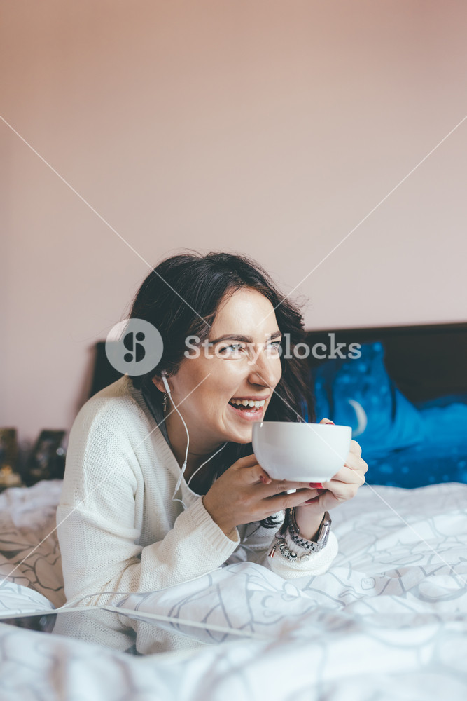 Young woman relaxing in her bedroom, enjoying coffee or tea and listening to music with earphones - serene concept
