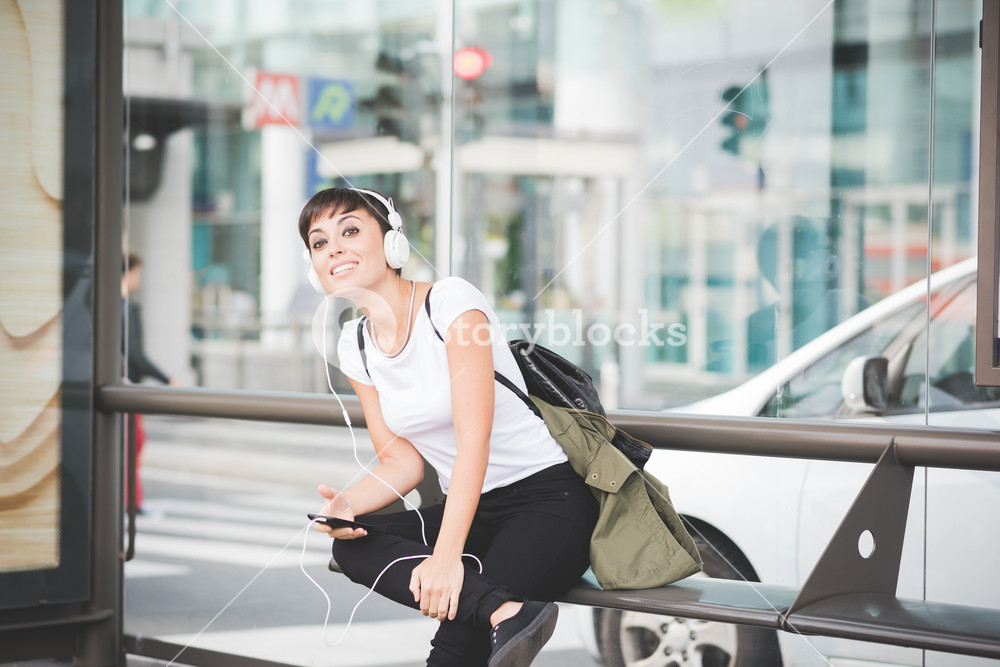 Young person sitting on a bench at a bus stop, enjoying music with headphones, looking to the side, using a smartphone - technology, social connection, music concept