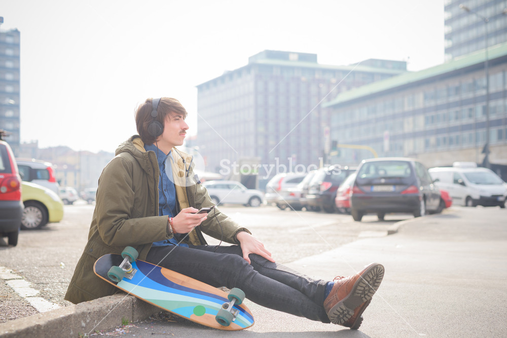 Young man skater seated on a sidewalk, enjoying music - relax, music concept - dressed in green coat and black jeans