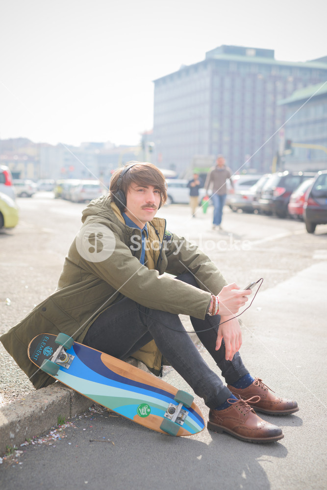 Young man skater seated on a sidewalk, enjoying music - relax, music concept - dressed in green coat and black jeans
