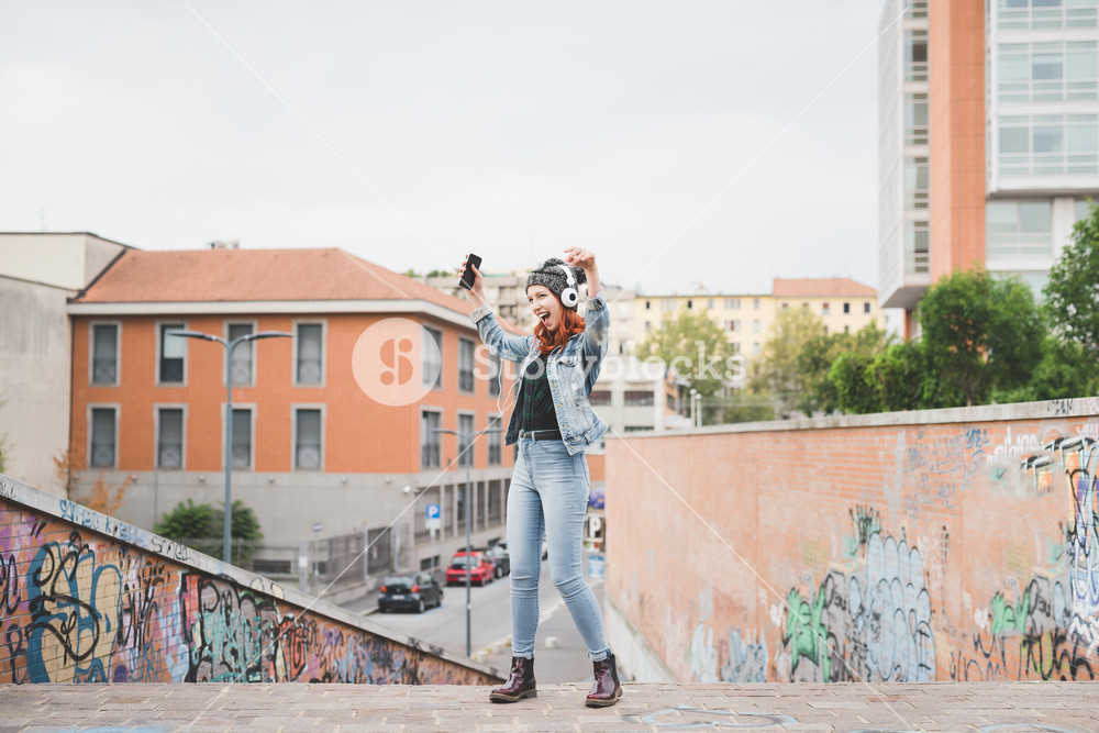 Young redhead woman dancing in the city, enjoying music with headphones and smartphone - music, relax, technology concept