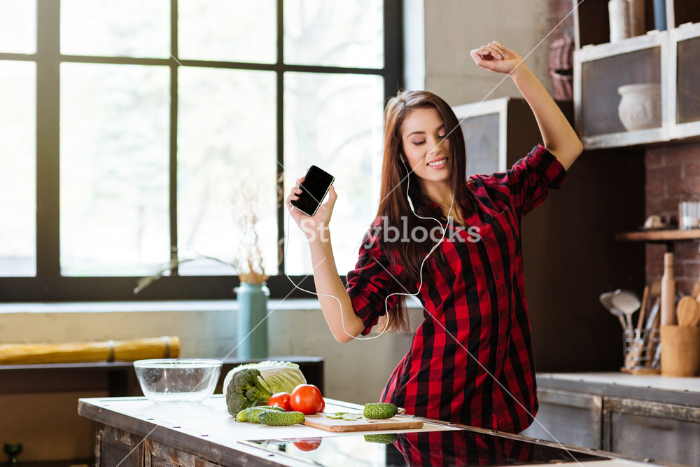 Woman in red shirt enjoying music, dancing in kitchen with phone and headphones.