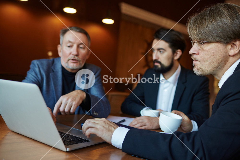 Profile view of concentrated middle-aged businessman in eyeglasses holding cup of coffee in hand while looking at computer screen and listening to his colleague with interest, waist-up portrait