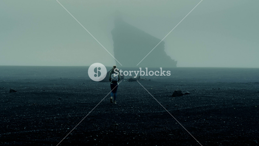 Woman Explorer Wearing A Beanie And Backpack Walk On Black Sand Beach In The Fog In The Iceland Wilderness With Monolith Rock In The Distance Slow Close Drone Shot 8 K