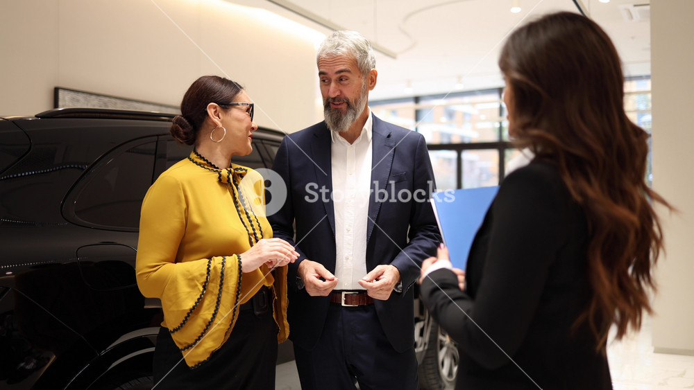 Senior man and woman talking to saleswoman about buying a new electric car in a modern vehicle dealership showroom