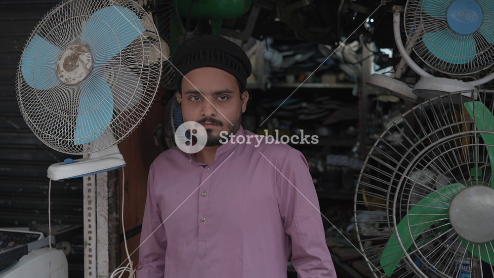A daylight outdoor shot of a young muslim Indian Asian man or male local business owner wearing traditional clothes, standing in front of the electric fan repair shop, looking at or facing the camera