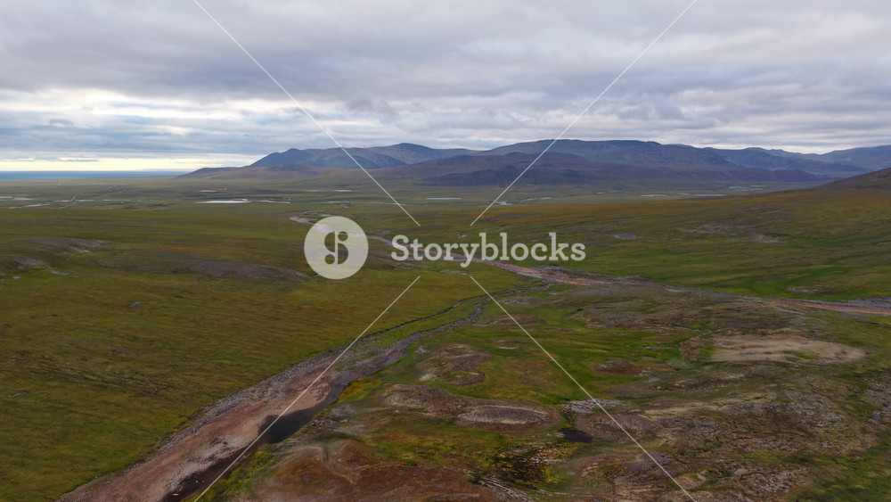 Rounded mounds and a dark creek mark frost heave terrain near Lavrentiya. Exposed gravel, lichen and wetland pools show Arctic geology