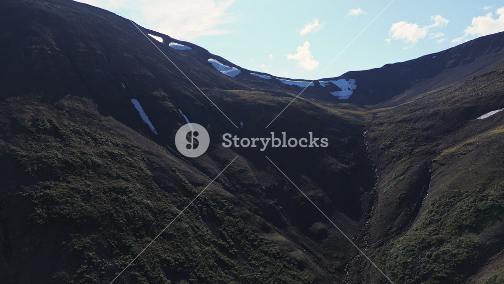Aerial view follows a high cirque on the Putorana Plateau where late summer snow lingers on dark slope. Steep wall, narrow ravine and alpine