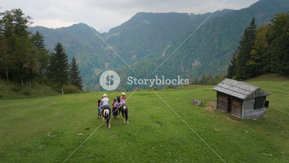 Couple enjoying a horseback ride across a green meadow, guided by instructors, surrounded by majestic mountain views and the peacefulness of nature, aerial shot.