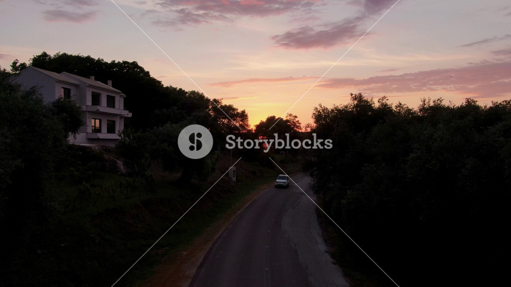 Beautiful aerial drone shot rising to reveal a stunning sunset over the rural hills and olive groves of Corfu, Greece, as a van drives on a winding road