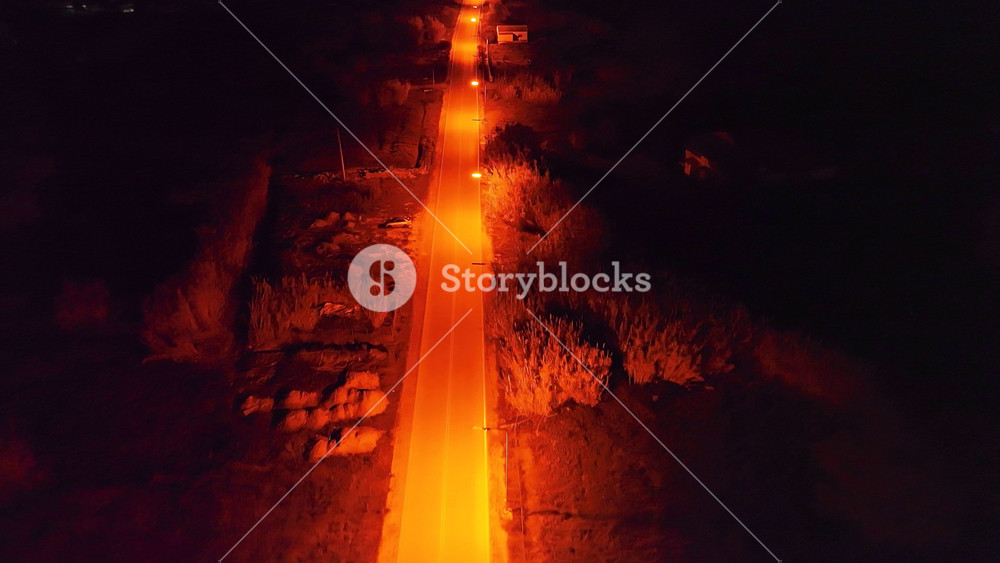 Eerie top-down aerial drone shot flying along a narrow, orange-lit road at night. A car's headlights approach in the darkness