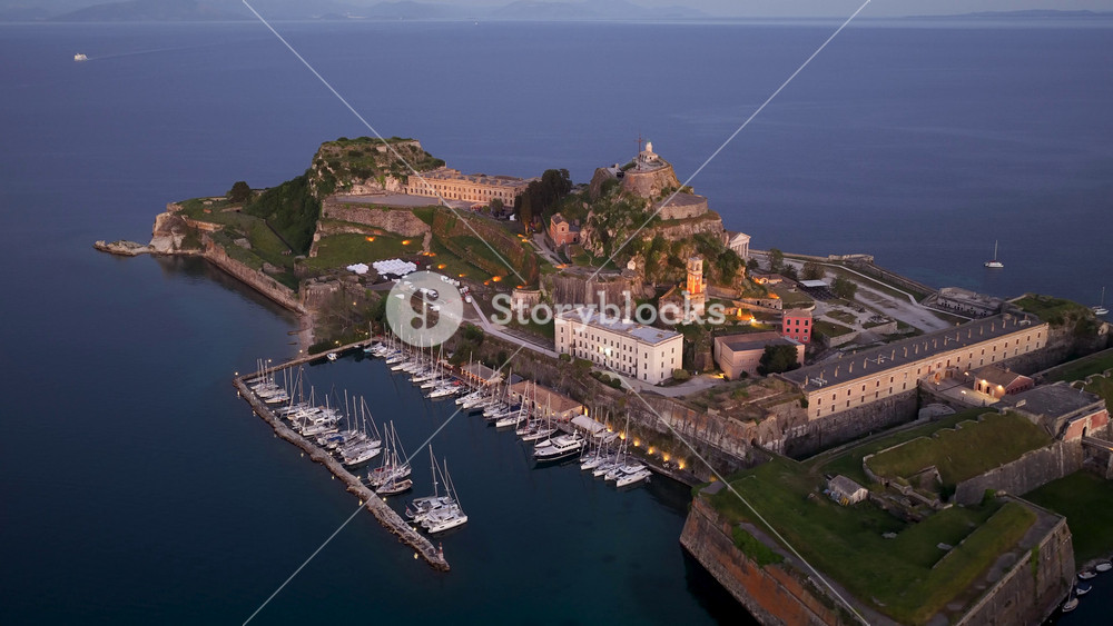 Aerial view of the Old Fortress in Corfu Town at twilight, featuring the historic Venetian citadel, illuminated architecture, and docked sailboats in the marina on the Ionian Sea