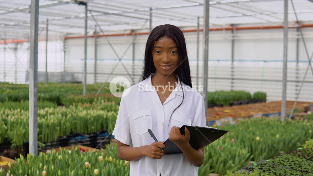 African American young girl florist or nerd in a white shirt stands on a background of rows of tulip seedlings with a pen and a tablet