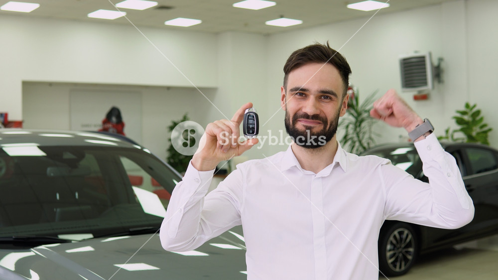 Emotional handsome guy raising hand up and smiling at camera, showing key near his brand new gray car. Rich turkish man buying auto at