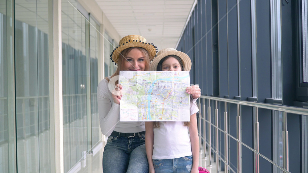 Beautiful woman with little girl holding a tourist map at the airport. Woman with daughter covers faces with tourist map