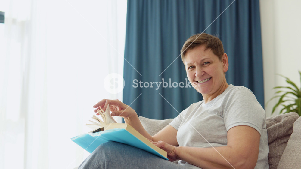 Relaxed senior woman sitting on sofa and reading book indoors at home. Leisure activity of mature female at comfort modern flat. Casual