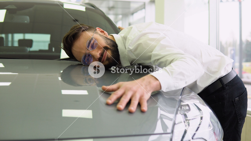 Happy Man Hugging Hood Of His New Car, Joyful Smiling Millennial Guy Dreaming About Buying Vehicle In Auto Showroom, Leaning At Surface Of
