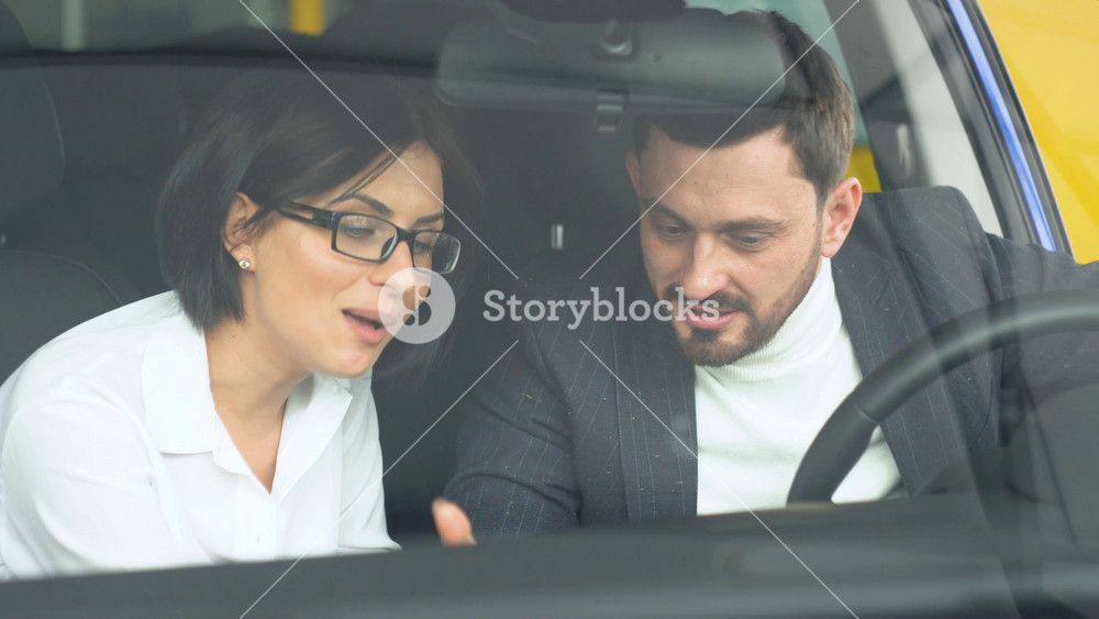 Stylish businessman sitting in car salon and talking with female car dealer. Young confident guy choosing automobile in showroom