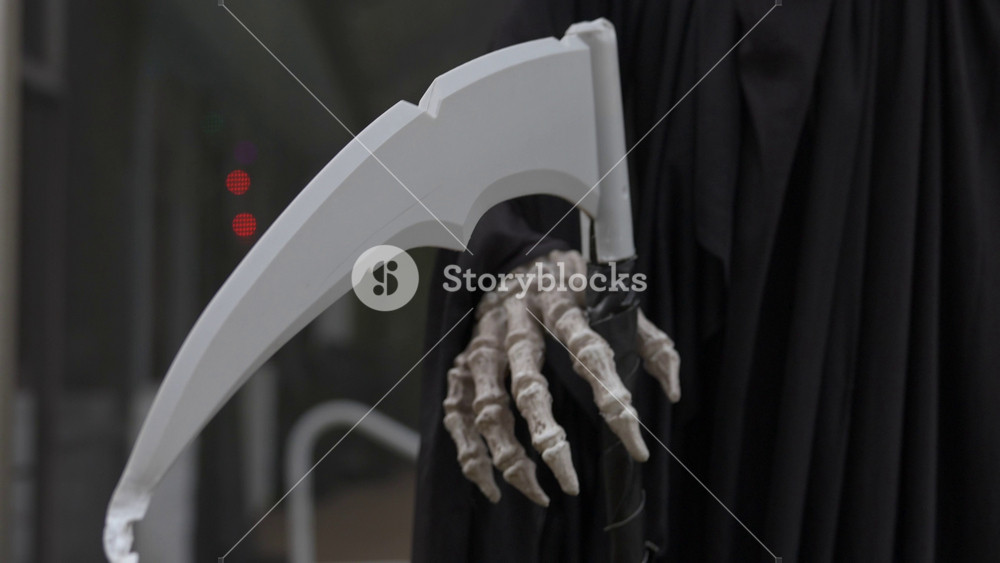 Close-up of a hooded grim reaper Halloween prop with a bony skeleton hand gripping a white scythe. Spooky outdoor decoration with a black