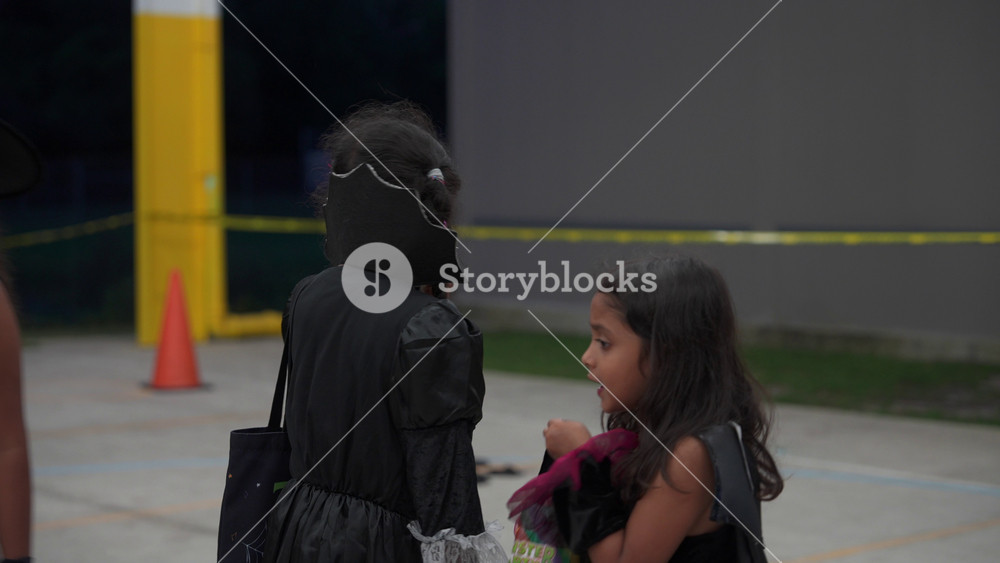 Orlando, Florida, USA - 10. 30. 2024: Two costumed girls walk together at a school Halloween event, one in a witch hat and black dress. They