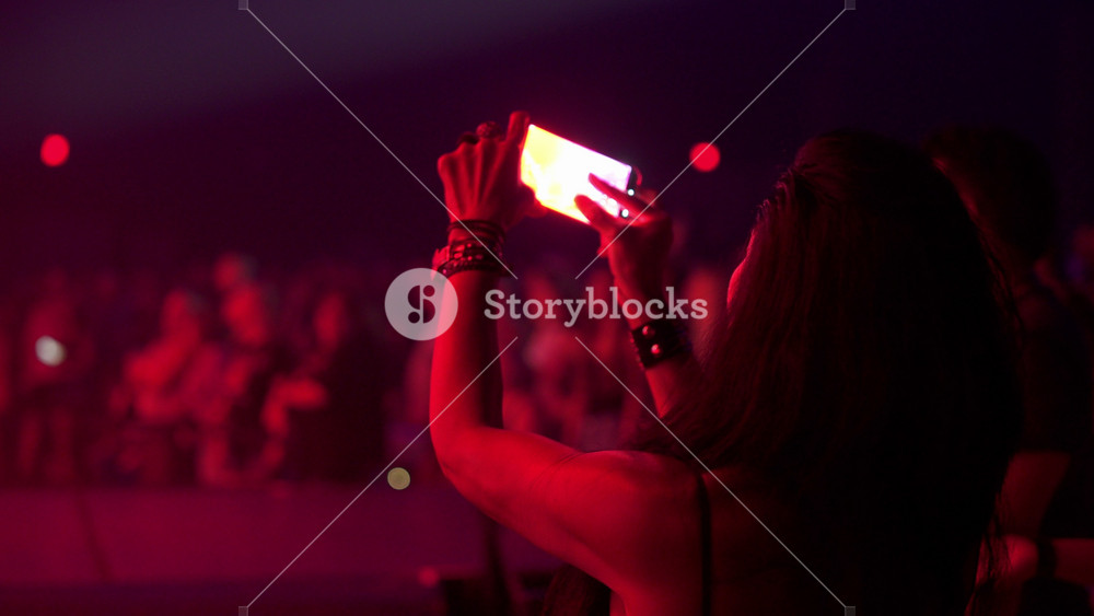 A concertgoer films the show with a phone under purple stage lights. The crowd stands close to the stage in a smoky indoor venue.