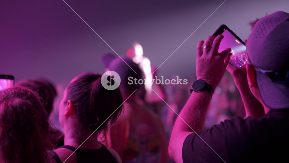 Close up of a concertgoer holding a smartphone to film a live performance in an indoor venue. Blue and purple lighting and haze create a