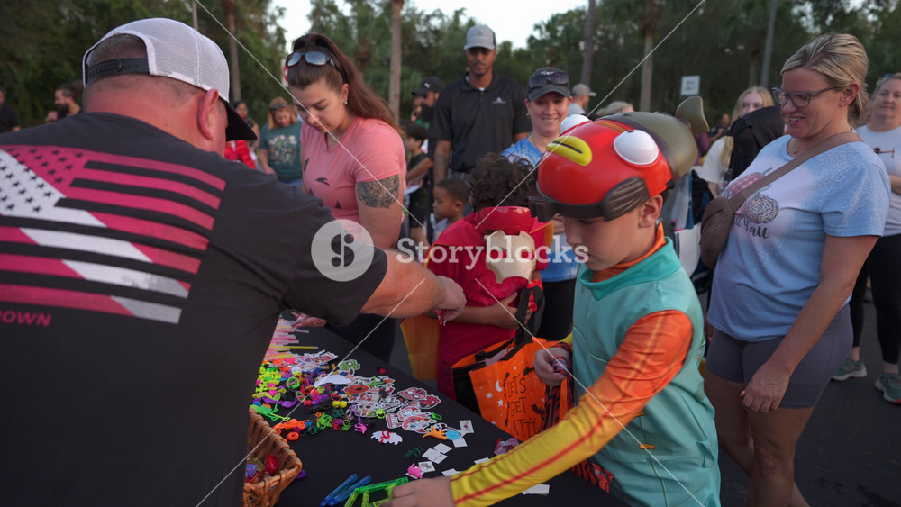 Orlando, Florida, USA - 10. 30. 2024: Kids in varied costumes reach for stickers and toys at a decorated table during a school event. Families
