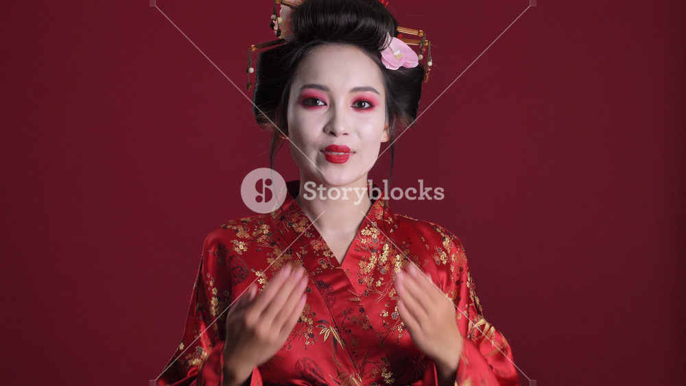 A lovely smiling young geisha woman in traditional japanese kimono is sending an air kiss to the camera isolated over red background