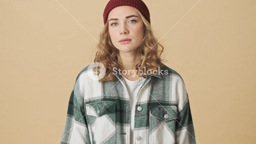 Calm pretty woman in knit hat and shirt becoming happy and showing thumb up after that over beige background