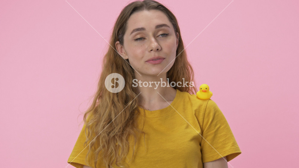 A cute young woman catches sight of with a small toy of yellow duck on her shoulder isolated over pink background
