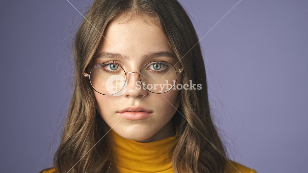 A close-up view of calm teenage girl wearing glasses is opening her eyes and looking to the camera standing isolated over a purple background in studio