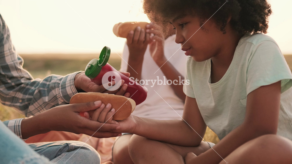 A close-up view of an afro-american mother and her children are eating hot-dogs while using ketchup sauce during a picnic outside at nature