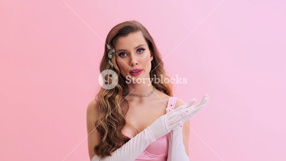 A displeased young woman wearing gorgeous evening pink dress with gloves and hairpins is clapping her hands standing isolated over a pink wall in the studio
