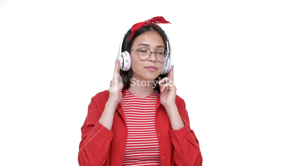 Happy young brunette girl in red shirt listening music on wireless earphones and dancing over white background isolated