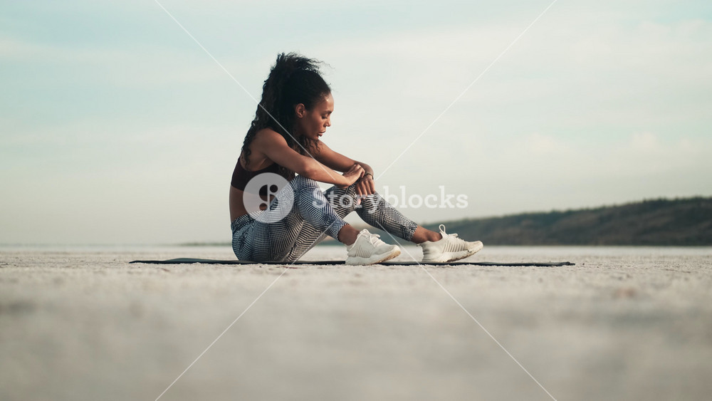 A focused african american woman wearing sportswear is using her fitness tracker sitting on the yoga mat outside at the beach