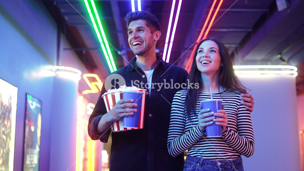 Smiling couple man and woman walking to the cinema holding a popcorn dating together inside the neon hall