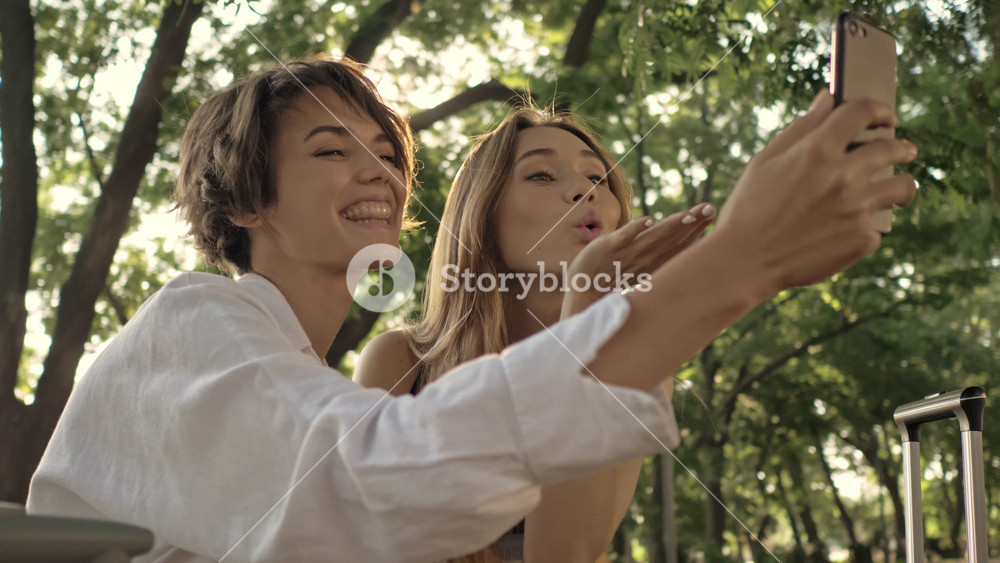 Two happy young tourist girls are sending an air kiss to the camera when having a video call in the city park