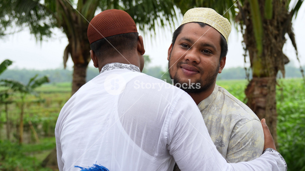 Bogura, Bangladesh - 06 February 2024: View of two Muslim men wearing traditional caps and panjabi hugging each other in a rural field with palm trees.